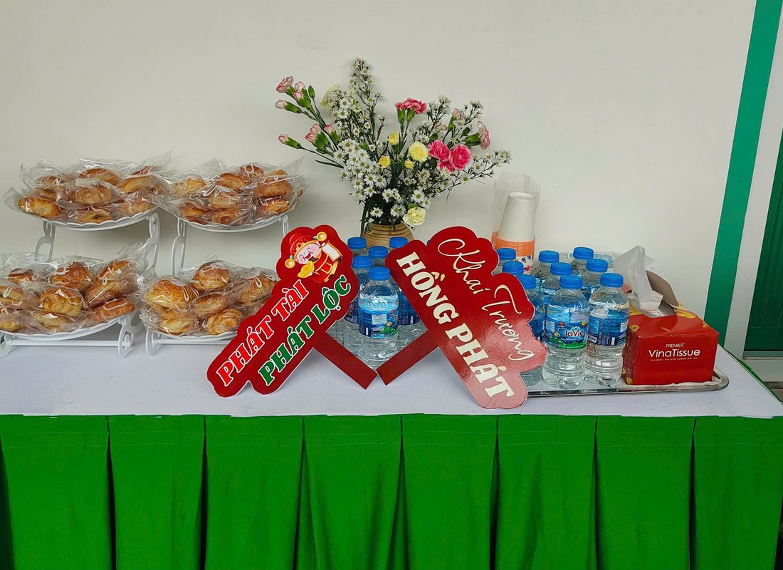 Bàn phủ khăn xanh với bánh đóng gói, nước uống và hoa trang trí.
