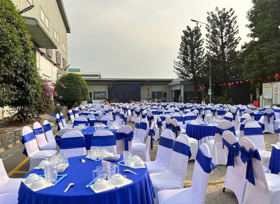 Bàn tròn phủ xanh và ghế trắng nơ xanh tại tiệc tất niên Nhật Tiến trong khuôn viên nhà máy.